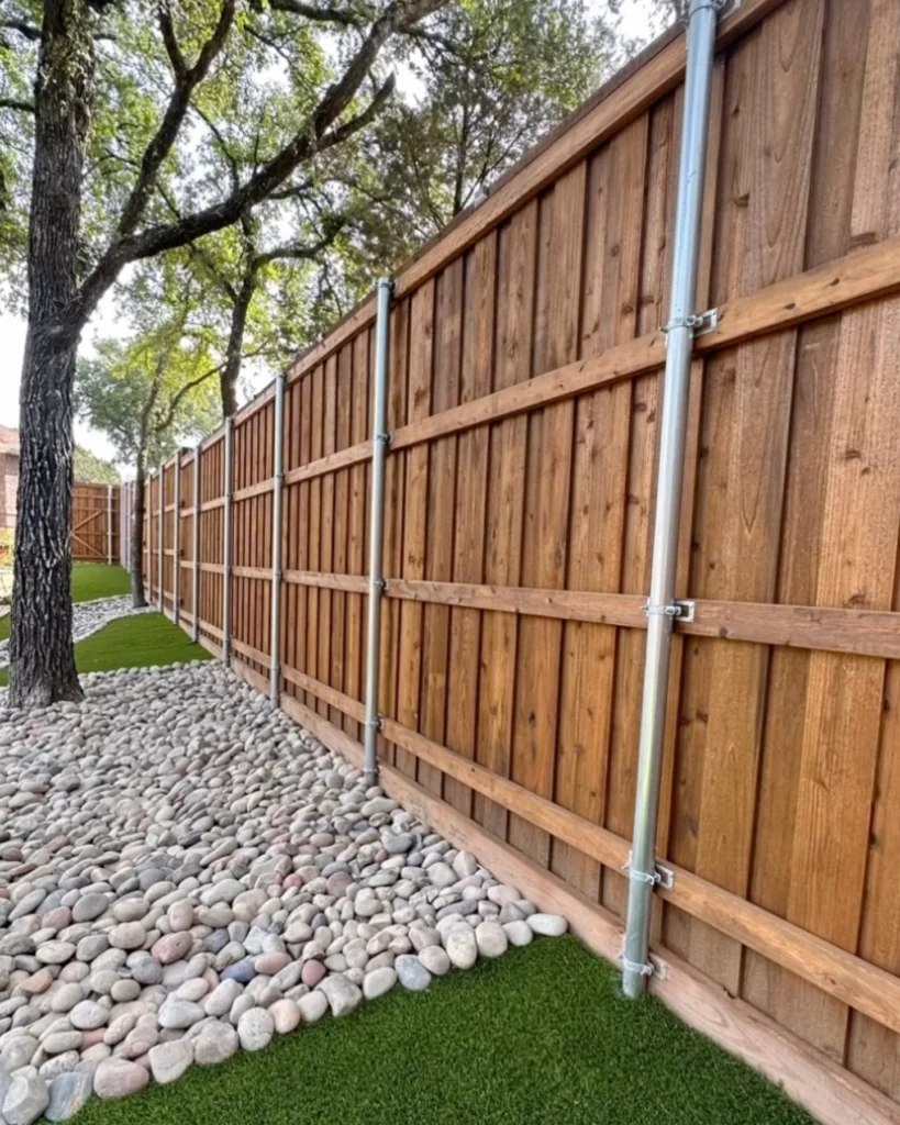 Cedar privacy fence on steel posts above a river-rock border and artificial turf