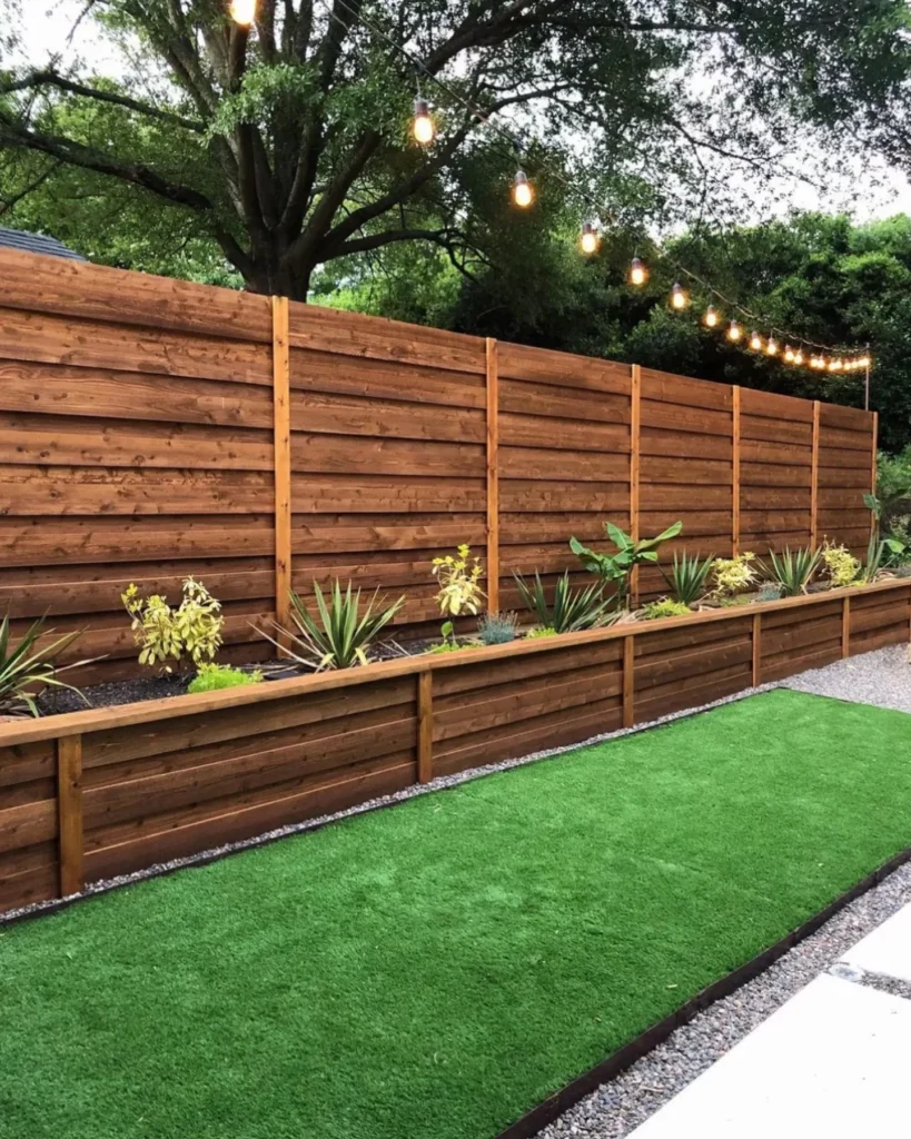 Horizontal cedar fence behind a raised planter bed with string lights over turf