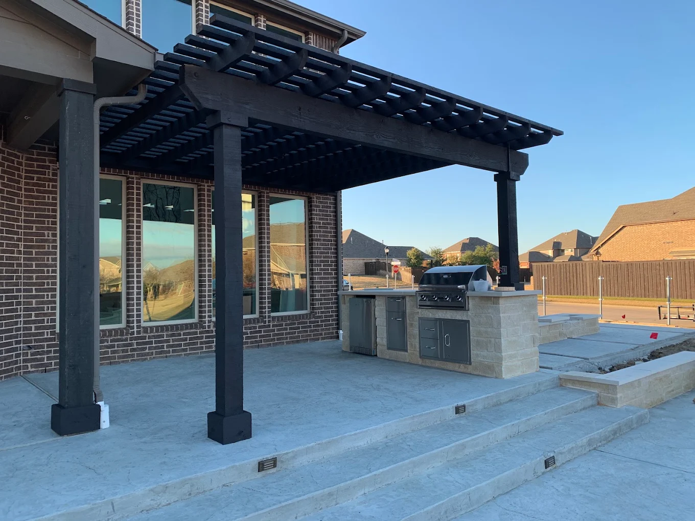 Black open-slat pergola attached to a brick home over an outdoor kitchen and concrete steps