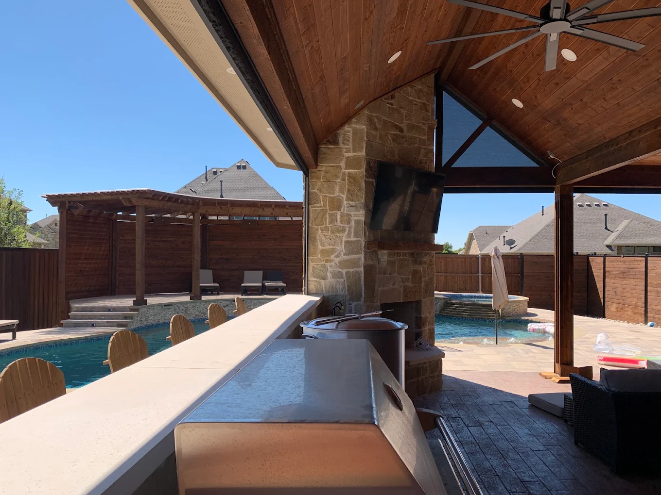 Gable patio cover with a stone fireplace, mounted TV and kitchen bar looking out over a pool
