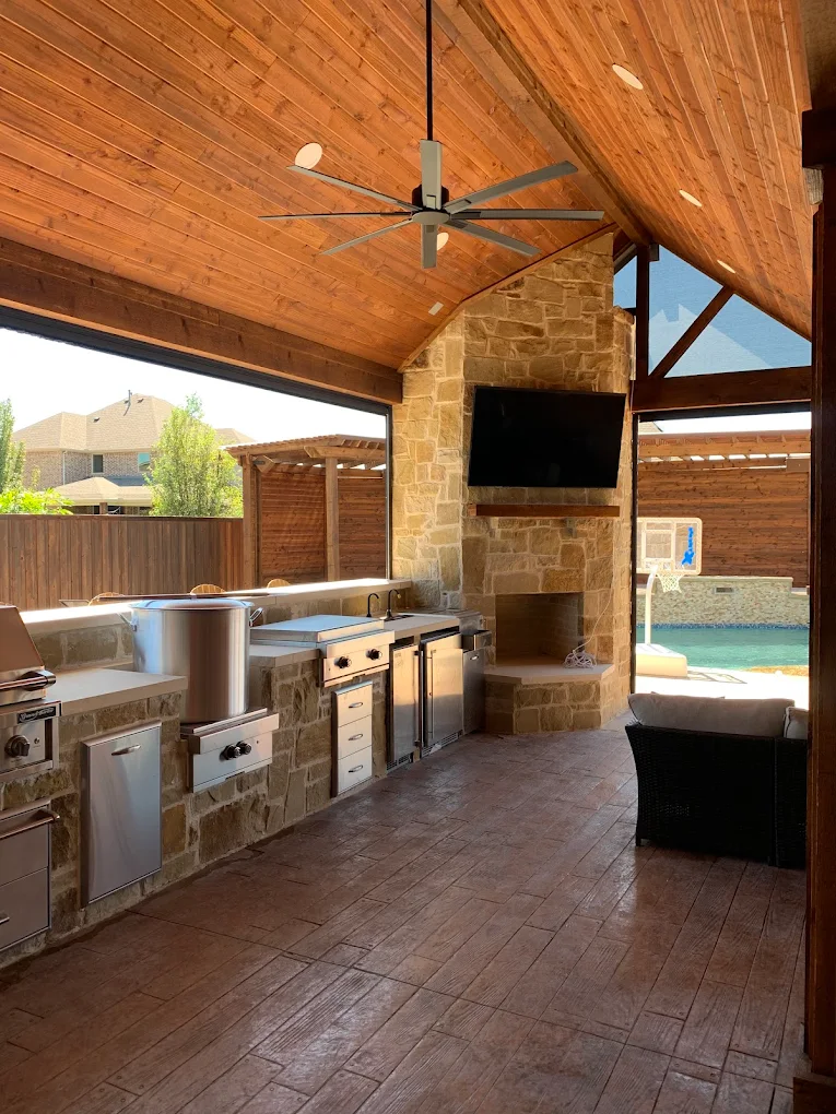 Vaulted cedar patio cover ceiling with a ceiling fan, stone fireplace, TV and outdoor kitchen beside a pool